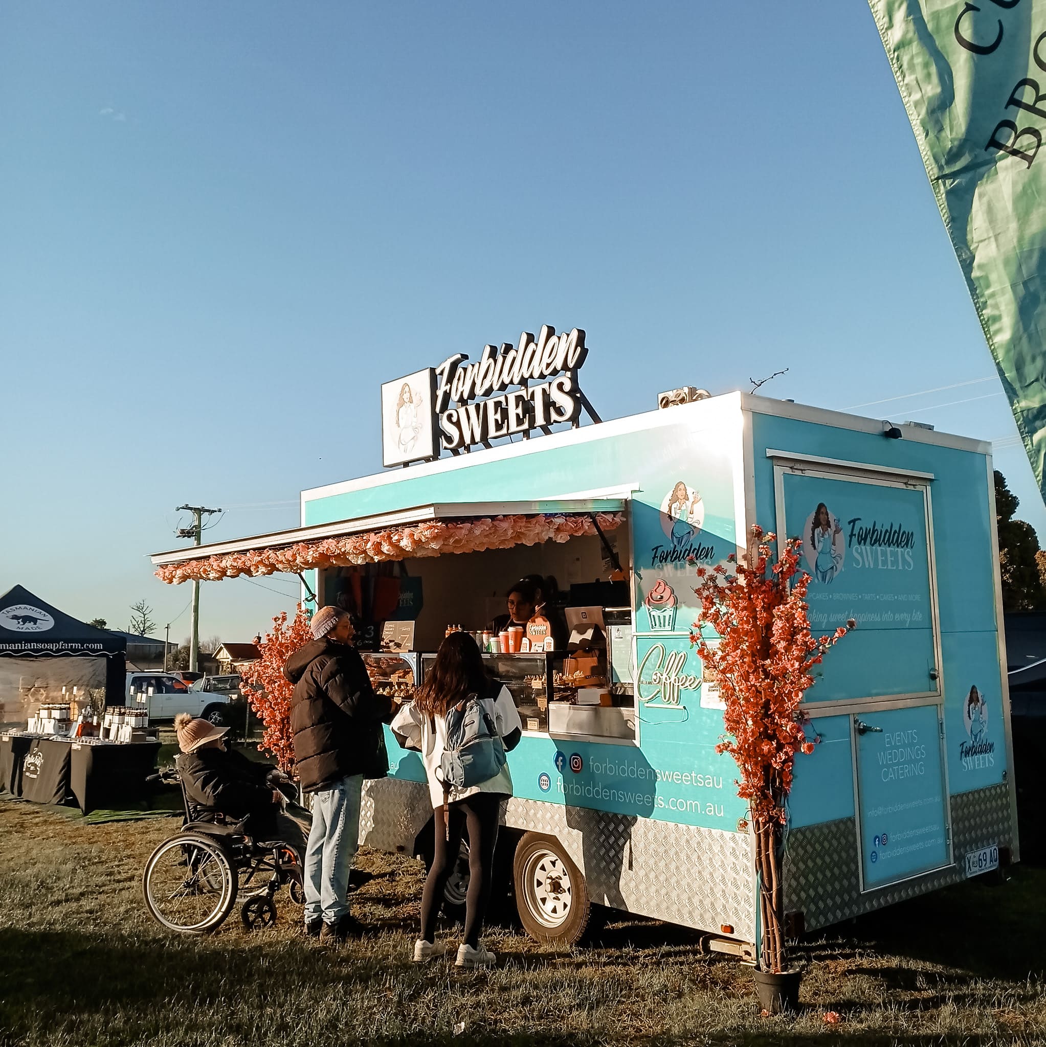 A turquoise food truck from Forbidden Sweets serving customers at the Wynyard Foreshore Market, with people lined up in front and stalls set up in the background on a sunny day. Ask ChatGPT