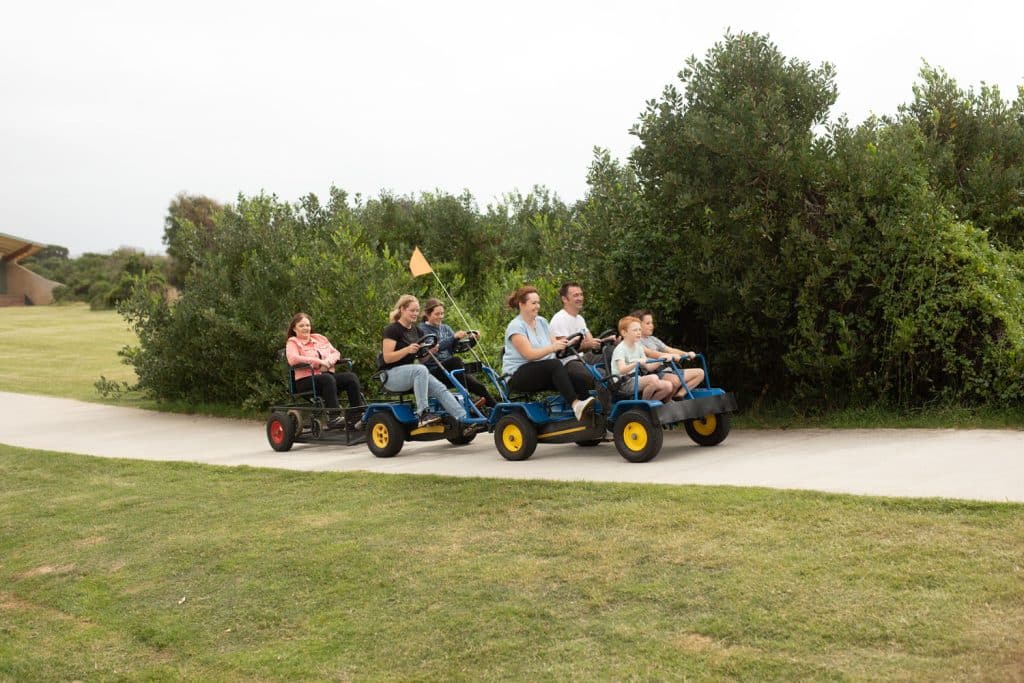A family smiling and enjoying a ride on a Pedal Buggies hire. A trailer is connected to the back towing a smiling woman in a wheelchair.