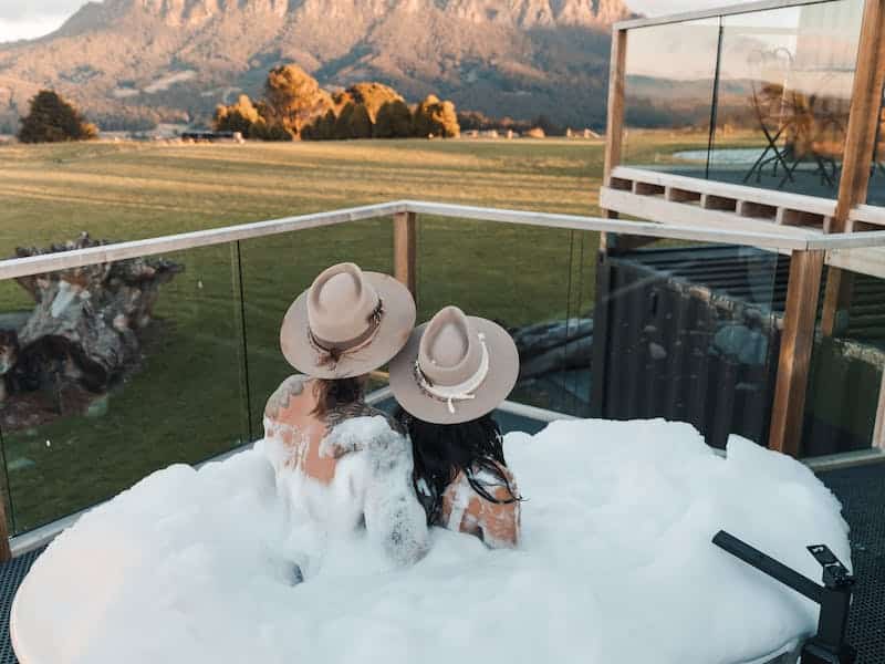 Two friends enjoy an outdoor bubble spa bath at Eagles Nest Retreat. The sun is shining across a field with mountains in the distance.