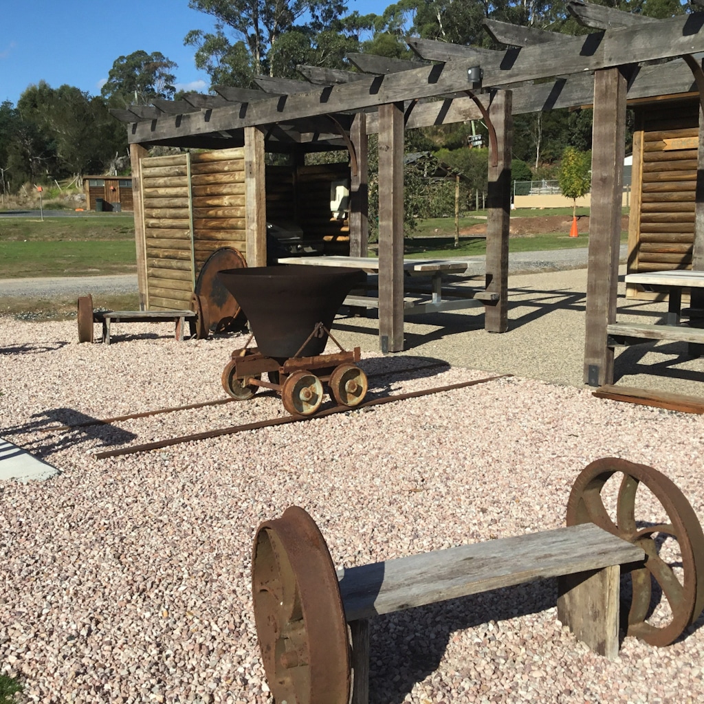 Rustic timber seating with rusted old machinery parts stand on gravel beside timber picnic tables at Lakeside Caravan and Camping ground in North West Tasmania