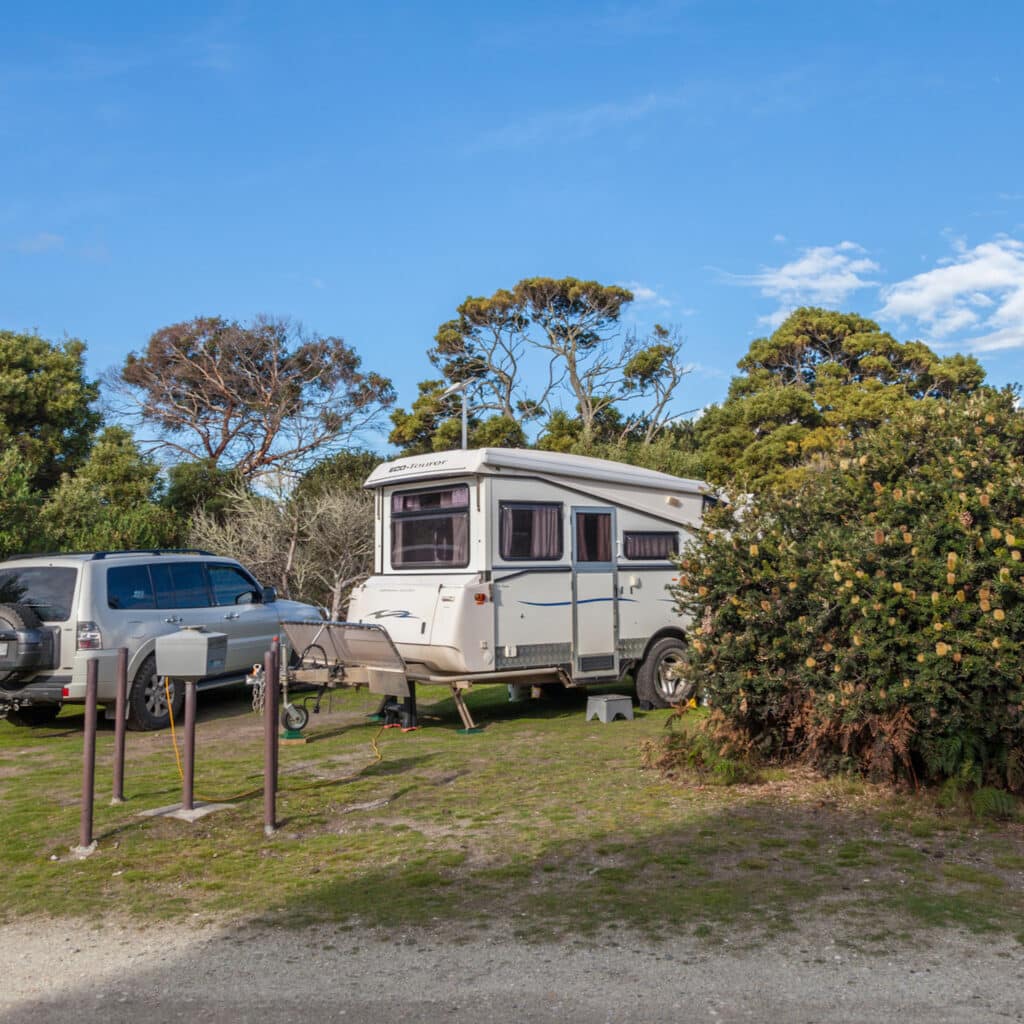 North West Tasmania caravan and camping adventures | A caravan is parked beside bushes with yellow flowers at a camping spot at Bakers Point Campground in North West Tasmania. On the left side of the caravan is parked a silver 4wd.