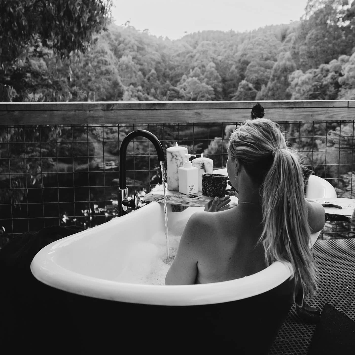 Woman with a blonde ponytail sits in an outdoor bath at Gleneagle Tasmania. She is looking out over a forest landscape.