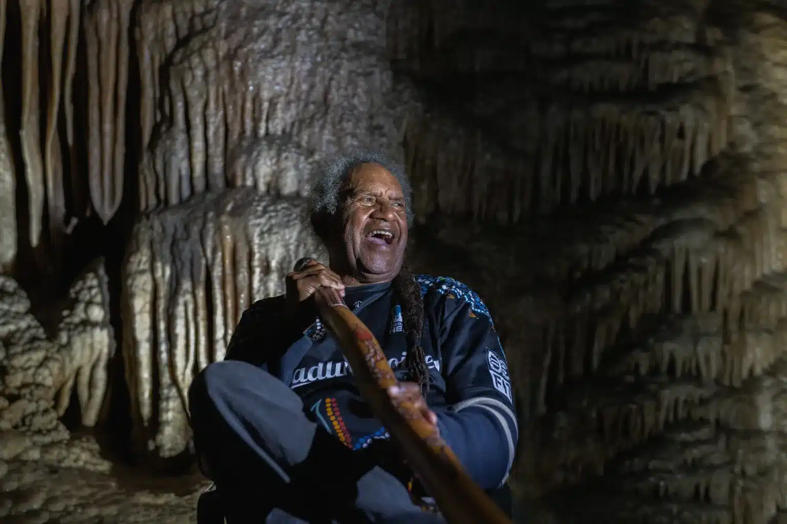 Aboriginal man smiling with didgeridoo inside limestone cave, surrounded by stalactites and natural rock formations at Gunns Plains Caves
