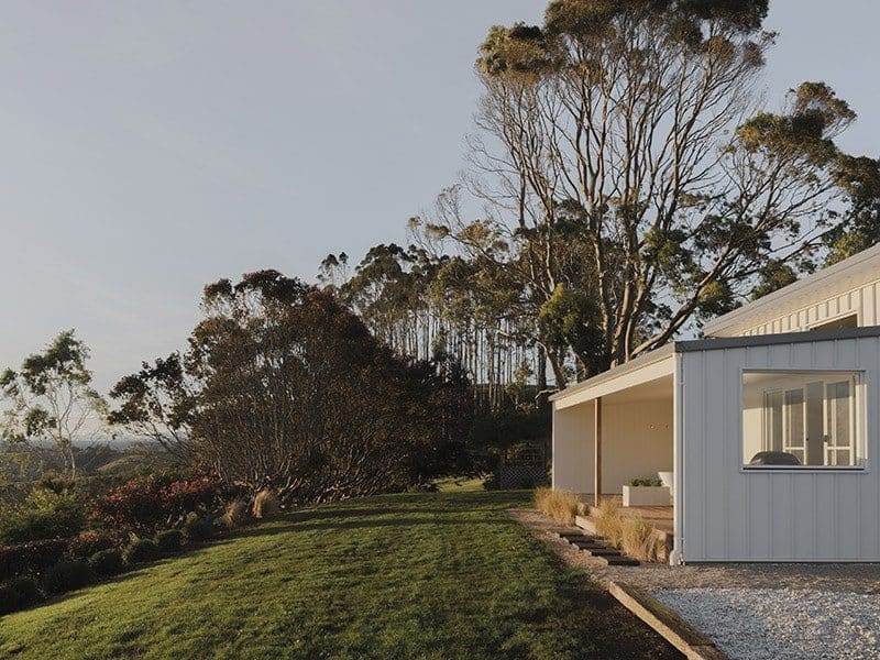 White cottage retreat with large windows and a covered patio, set on a grassy hillside surrounded by tall trees and native bushland.