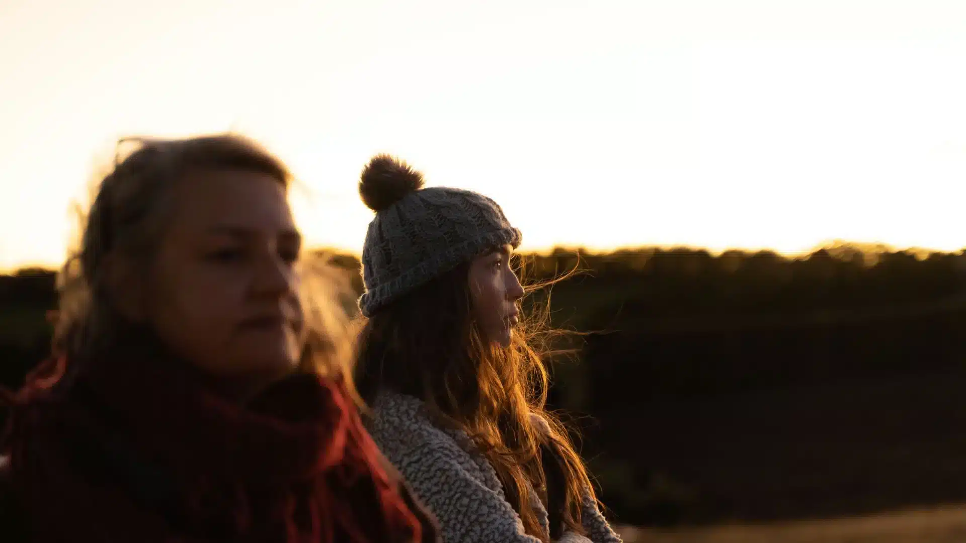Two women, one wearing a beanie at golden hour, looking across the landscape. The light is reflecting off their hair and the horizon. Light up the coast.