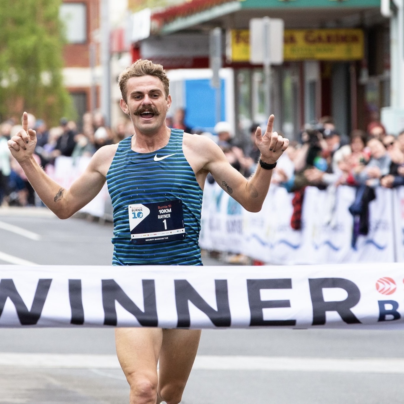 Runner crossing the finish line as the winner of the Burnie 10 road race in Tasmania, celebrating with arms raised as the crowd cheers.