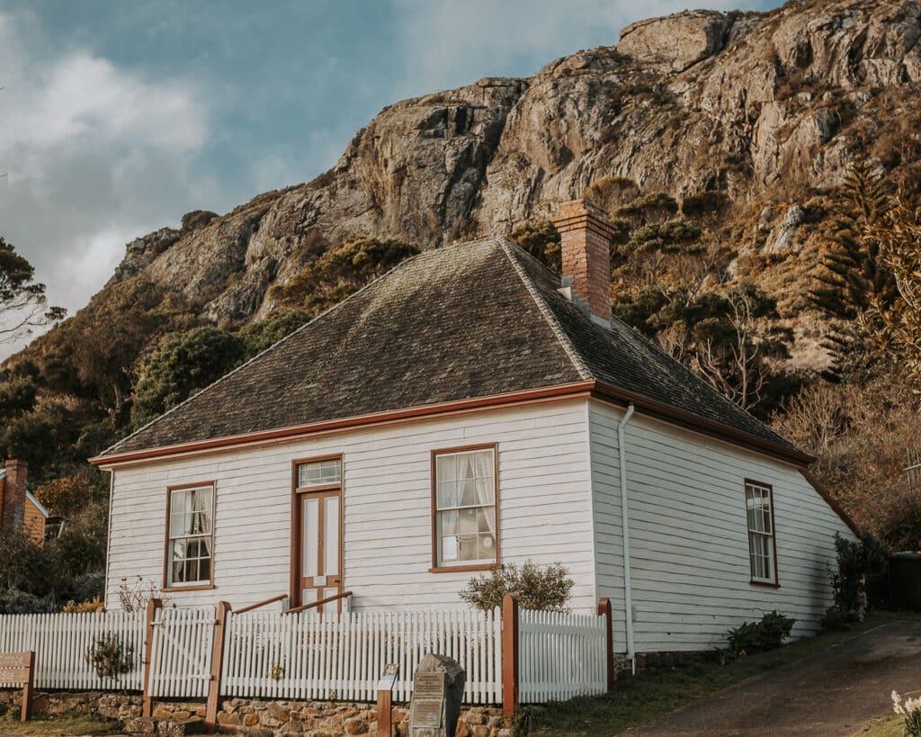 Historic white weatherboard cottage with brick chimney and dark slate roof nestled at the base of Stanley Nut's dramatic volcanic rock face. White picket fence borders the heritage building, with native vegetation covering the steep cliffs behind.