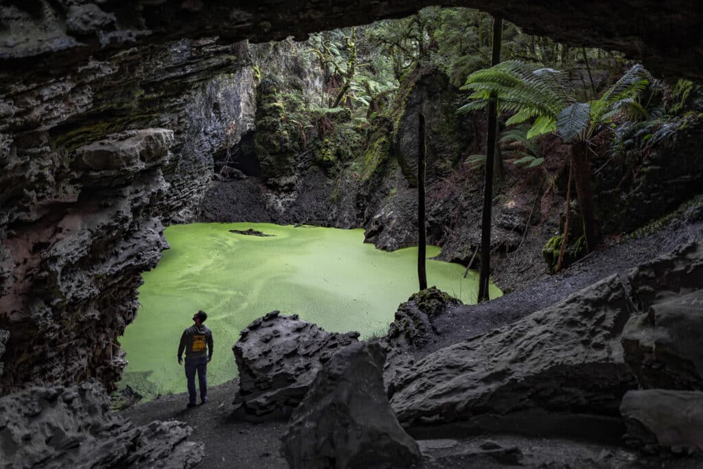 Person standing inside Trowutta Arch looking out at a luminous green sinkhole pool surrounded by dark volcanic rock. Tree ferns and rainforest vegetation frame the collapsed cave opening, with moss-covered rocks scattered around the water's edge.