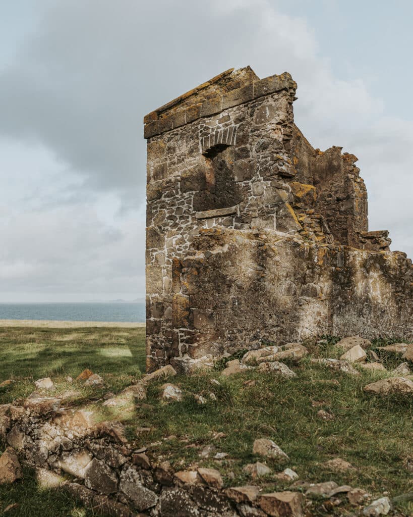 Weathered stone ruins of a historic building stand alone in coastal grassland with Bass Strait visible in the background. The crumbling structure shows lichen-covered stonework and empty window frames, with scattered rocks at its base under overcast skies.