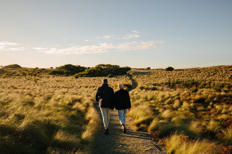 Two people walking away along a narrow track through coastal grassland in soft afternoon light.
