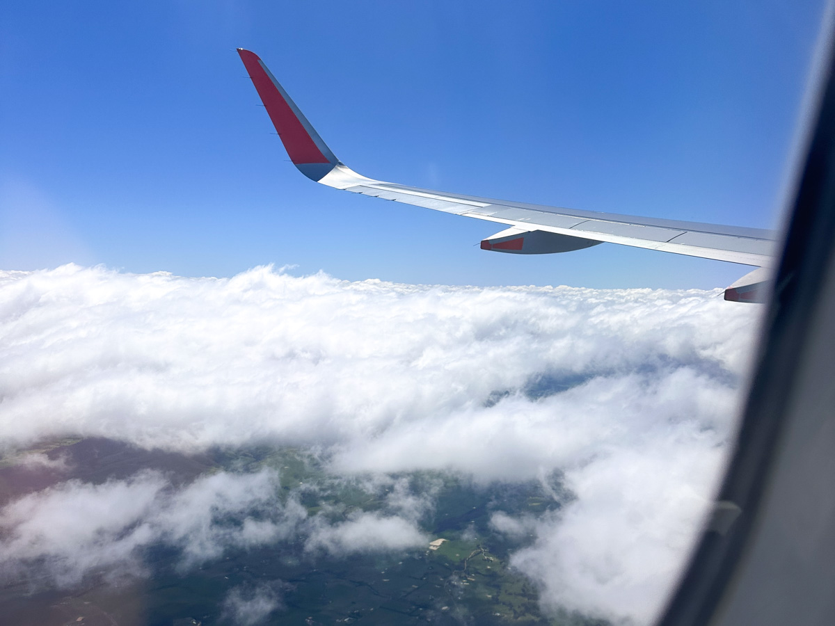 View from a plane window of a wing with a red winglet above a thick layer of white cloud, with a gap showing green patchwork farmland below and clear blue sky above.