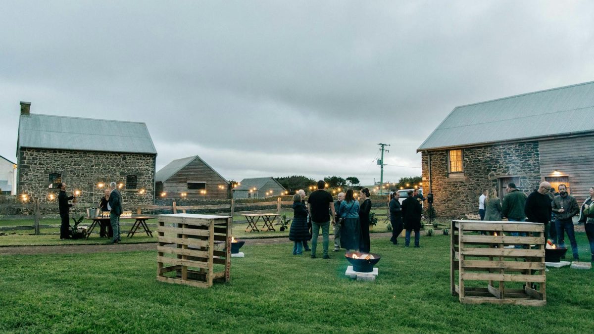 Guests gather around fire bowls and festoon lights between old stone farm buildings at dusk, Forage Festival, North West Tasmania