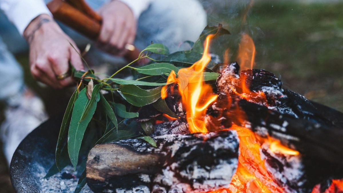 Hands holding gum leaves over a fire in a metal fire bowl, with smoke and flames rising